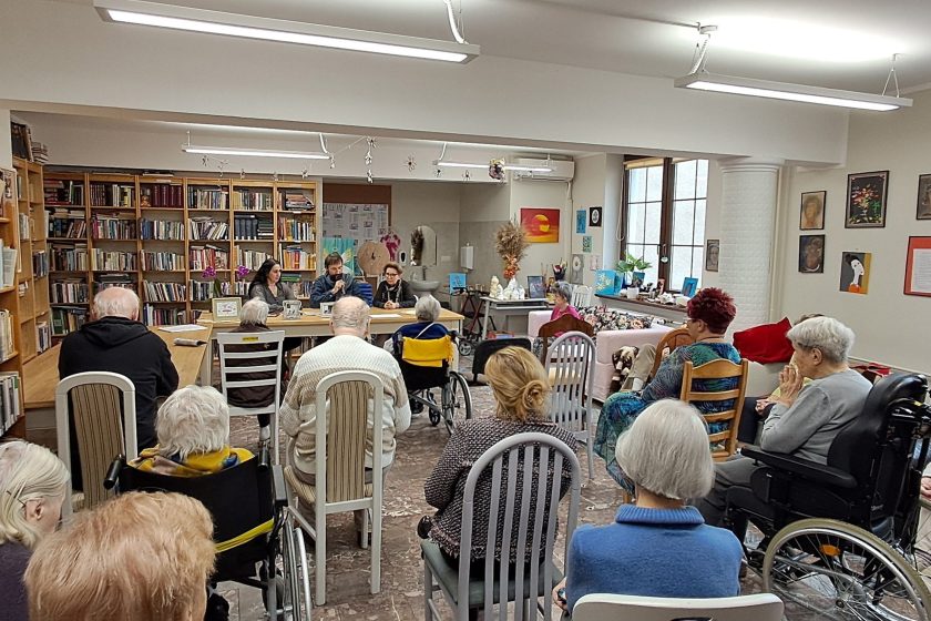 Ujęcie z tyłu, ukazujące salę z biblioteką i grupą seniorów siedzących przodem do stołu prelegentów. Trójka prowadzących siedzi za stołem – dwoje z nich mówi do mikrofonu. W tle regały z książkami, dekoracje ścienne oraz wystrój nadający przestrzeni ciepła i kameralnego charakteru. Publiczność uważnie słucha