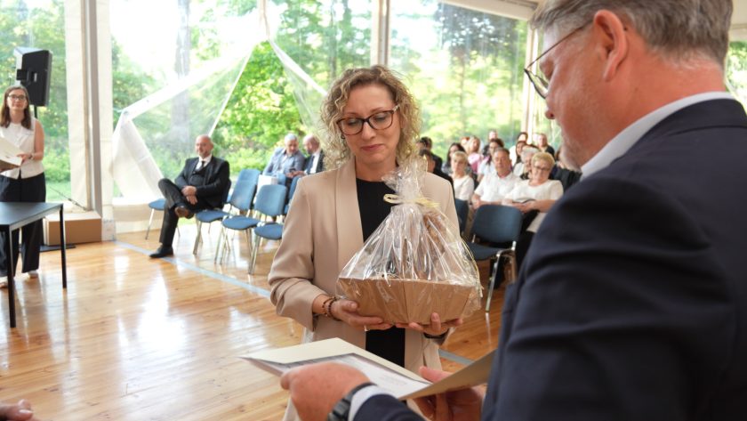 Kobieta w jasnym żakiecie i okularach odbiera pamiątkowy kosz upominkowy. Trzyma go obiema rękami, a mężczyzna w garniturze wręcza jej dokument. W tle widoczna grupa uczestników uroczystości oraz dekoracje wnętrza.