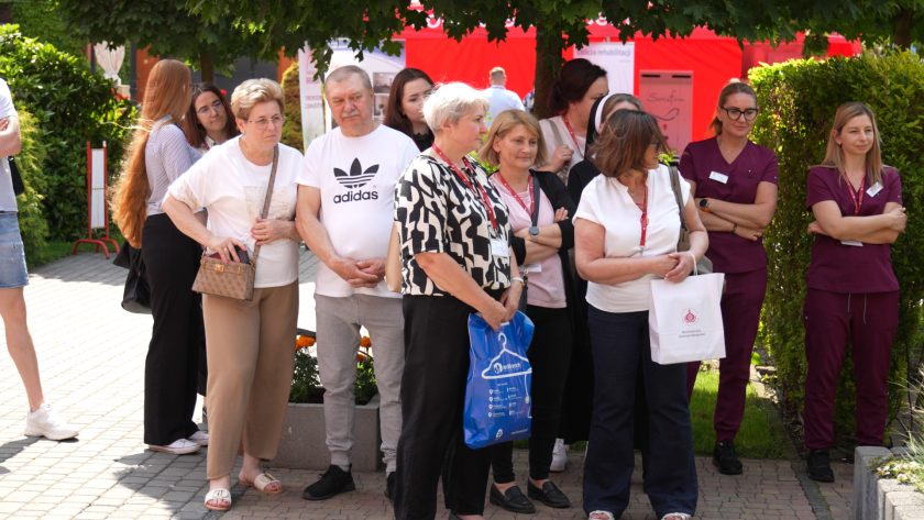 Uczestnicy wydarzenia stojący w cieniu drzewa, uważnie obserwują pokaz rehabilitacyjny.