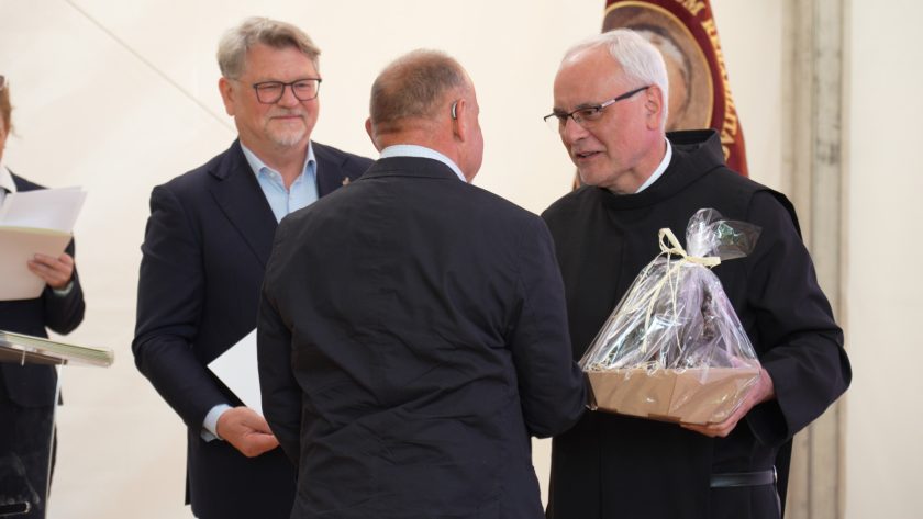 Mężczyzna w garniturze i bonifrater w habicie uściskują dłonie podczas wręczenia pamiątkowego kosza. Uśmiechnięty brat Franciszek Salezy Chmiel trzyma prezent, a za nimi stoi sztandar. W tle widoczna kobieta z dokumentami.