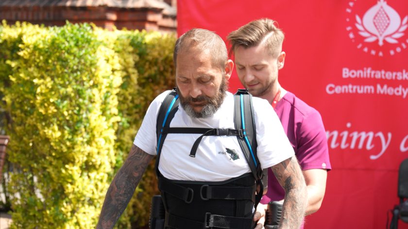 Zbliżenie na twarz skupionego pacjenta w egzoszkielecie oraz fizjoterapeuty pomagającego w ćwiczeniu chodu.