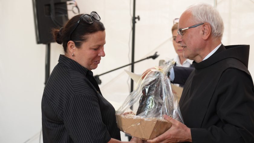 Brat bonifrater uroczyście wręcza prezent kobiecie w ciemnej bluzce w prążki. Ujęcie przedstawia moment przekazania pamiątkowego kosza owiniętego folią, w jasnym wnętrzu z obecnością mikrofonów i sprzętu nagłośnieniowego.