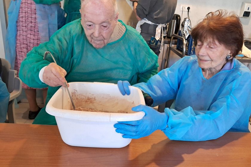 Starszy mężczyzna miesza w dużej misie czekoladową masę, a siedząca obok seniorka przytrzymuje pojemnik. Oboje mają na sobie niebiesko-zielone fartuchy ochronne i rękawiczki. Zdjęcie przedstawia zaangażowanie i współpracę podczas zajęć kulinarnych. W tle widoczne są elementy wyposażenia kuchennego