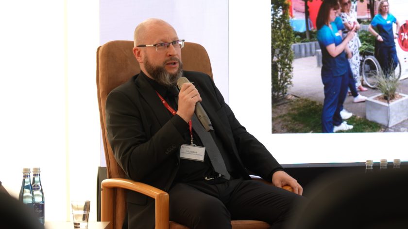 Jeden z panelistów – mężczyzna z mikrofonem – bierze udział w dyskusji podczas konferencji, siedząc na scenie.