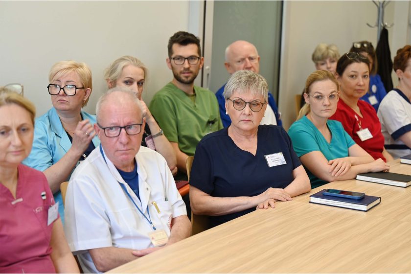 Grupa pracowników medycznych, kobiety i mężczyźni w różnokolorowych uniformach, siedzi przy stole konferencyjnym, słuchając z powagą wypowiedzi.