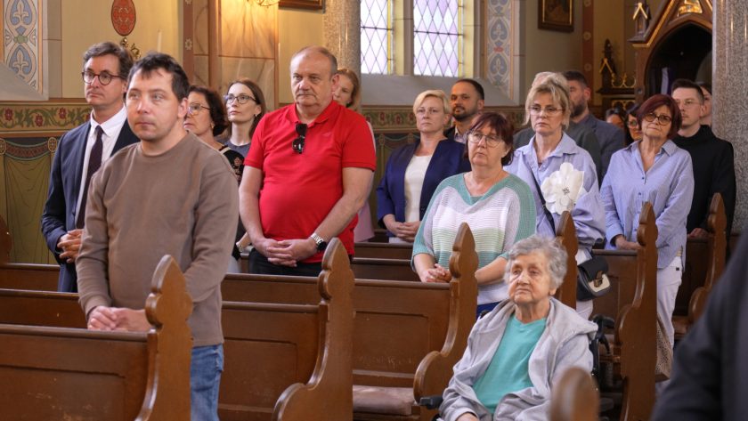 Grupa uczestników nabożeństwa stojących w ławkach kościelnych. Na pierwszym planie starsza kobieta na wózku inwalidzkim oraz osoby w różnym wieku w skupieniu biorące udział w liturgii. W tle widoczne zdobienia kościoła i światło wpadające przez witraże.