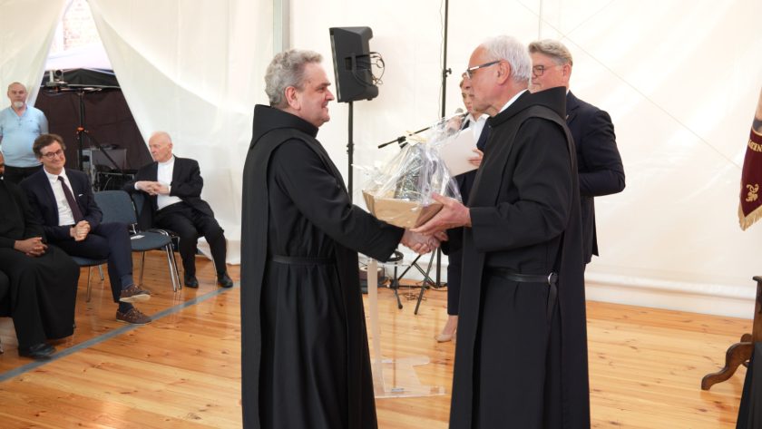 Dwóch zakonników bonifraterskich w habitach uściskuje sobie dłonie podczas wręczenia prezentu. Jeden z nich trzyma w rękach kosz upominkowy zapakowany w folię. W tle widoczni uczestnicy siedzący na krzesłach i sprzęt techniczny.