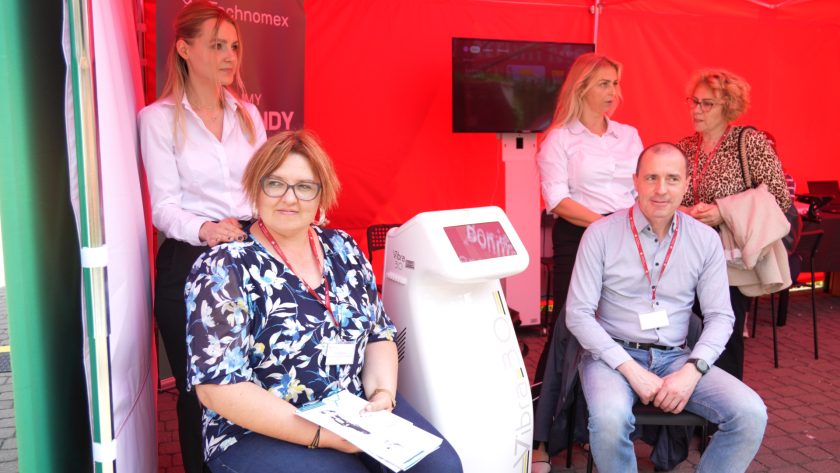 Kobieta i mężczyzna siedzą przy urządzeniu medycznym Vibra 3.0 w czerwonym namiocie wystawowym; w tle stoją trzy kobiety prowadzące rozmowę.
