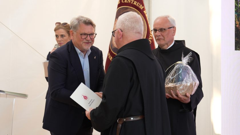 Mężczyzna w garniturze ściska dłoń zakonnika w habicie, przekazując mu dyplom. Obok drugi zakonnik trzyma pamiątkowy kosz upominkowy. W tle widoczny bordowy sztandar z wizerunkiem św. Jana Bożego.