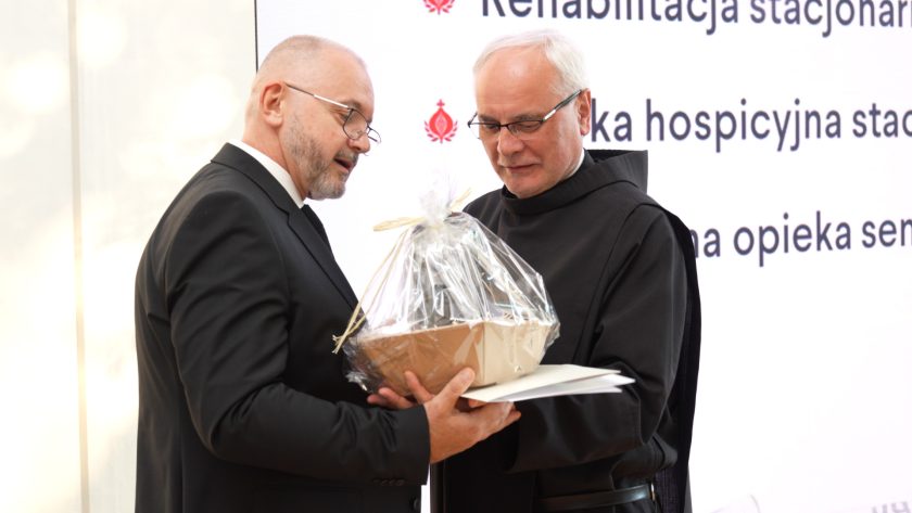 Zakonnik w habicie wręcza prezent mężczyźnie w garniturze. Obaj trzymają kosz upominkowy zawinięty w folię. W tle widoczna prezentacja multimedialna z hasłami o działalności Bonifraterskiego Centrum Medycznego.