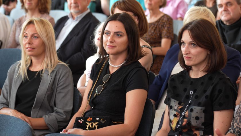 Trzy kobiety siedzą w pierwszym rzędzie i uważnie słuchają wystąpień. Dwie z nich mają długie, rozpuszczone włosy, a wszystkie ubrane są elegancko, w odcieniach czerni i szarości. W tle inne osoby śledzą przebieg wydarzenia.
