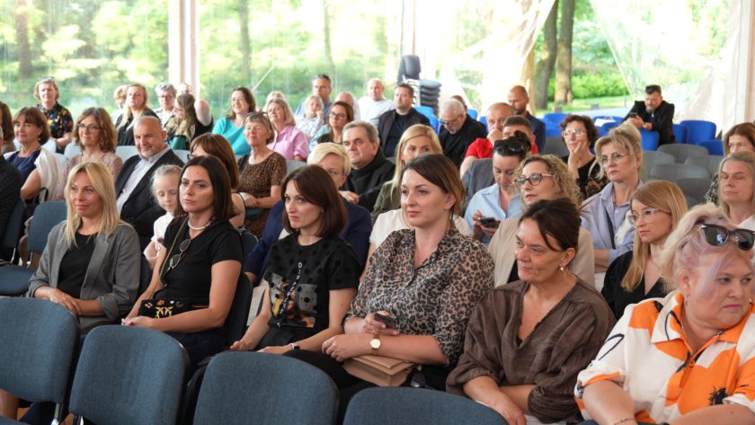 Szeroki kadr sali pokazujący licznych uczestników jubileuszu, siedzących w rzędach na tle przeszklonego namiotu i zieleni. Na pierwszym planie kobiety w różnym wieku, a w tle – mieszana grupa gości i pracowników.