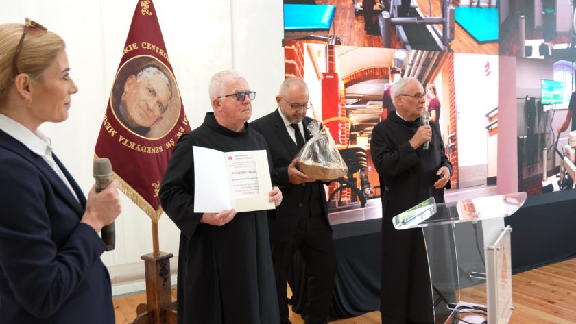 Brat Franciszek Salezy Chmiel trzyma otwarty dyplom z podziękowaniem, obok mężczyzna z koszem upominkowym, a trzeci zakonnik przemawia do mikrofonu. W tle plansza z grafiką szpitalnych wnętrz i kobieta z mikrofonem.