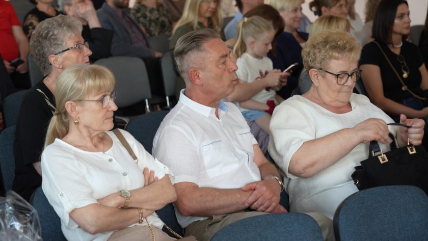 Grupa starszych osób i dzieci siedzących w rzędach – niektórzy słuchają uważnie, inni przeglądają torebki lub korzystają z telefonów. Na pierwszym planie kobieta w białej bluzce i mężczyzna w jasnej koszuli.