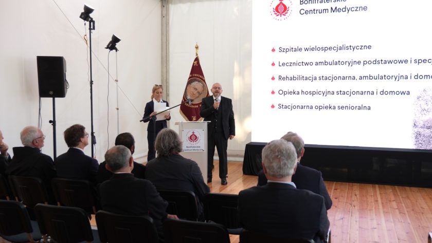 Prelegent w garniturze z mikrofonem stoi przy mównicy, a obok niego kobieta prowadząca odczyt. Za nimi duży ekran z listą zakresu działalności Bonifraterskiego Centrum Medycznego. W tle siedząca publiczność.