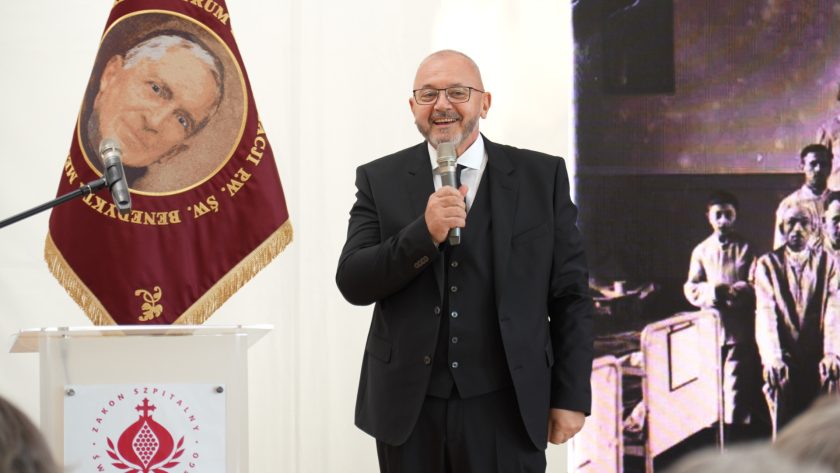 Ujęcie mężczyzny w ciemnym garniturze mówiącego do mikrofonu z uśmiechem, stojącego przed ekranem z historycznym zdjęciem szpitalnym. Po lewej stronie widoczna mównica z herbem Zakonu Bonifratrów i bordowy sztandar.