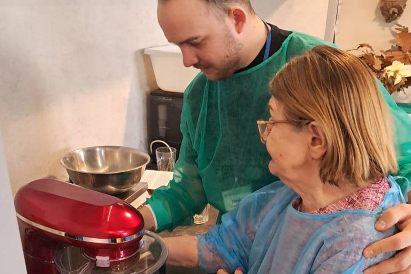 Młody opiekun asystuje starszej kobiecie przy obsłudze czerwonego robota kuchennego. Oboje skupieni, mają na sobie ochronne fartuchy. Mężczyzna delikatnie podtrzymuje seniorkę, wspierając ją w samodzielnym działaniu. W tle kuchenne blaty, miski i sprzęt AGD. Ciepła, wspierająca atmosfera międzypokoleniowej współpracy