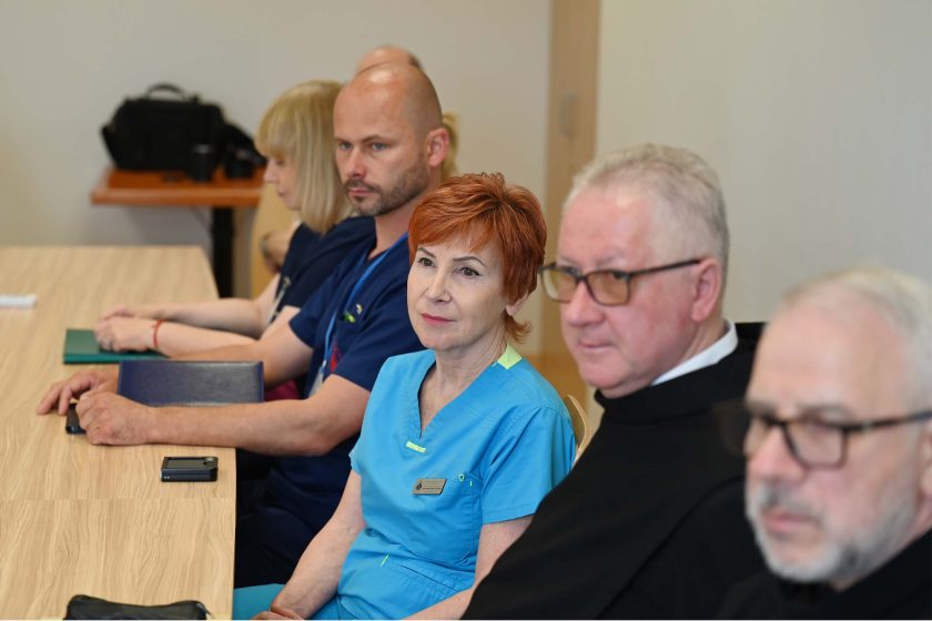Personel medyczny i zakonnicy siedzą przy stole, skupieni na rozmowie. W centrum kadru kobieta w niebieskim uniformie z rudymi włosami i brat zakonny w okularach.