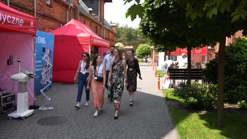 Grupa uczestników konferencji spaceruje alejką pomiędzy czerwonymi namiotami wystawienniczymi Bonifraterskiego Centrum Medycznego, otoczona ceglanymi zabudowaniami i zielenią.