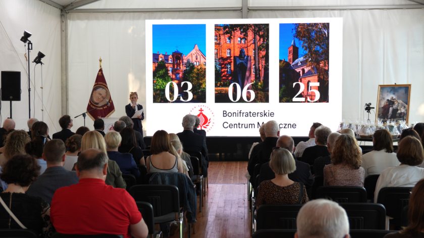 Widok od tyłu na uczestników siedzących na sali podczas inauguracji wydarzenia. Na scenie kobieta w ciemnym garniturze przemawia do mikrofonu, obok niej wyświetlony na ekranie napis „03 06 25 Bonifraterskie Centrum Medyczne” z trzema zdjęciami klasztoru i szpitala.