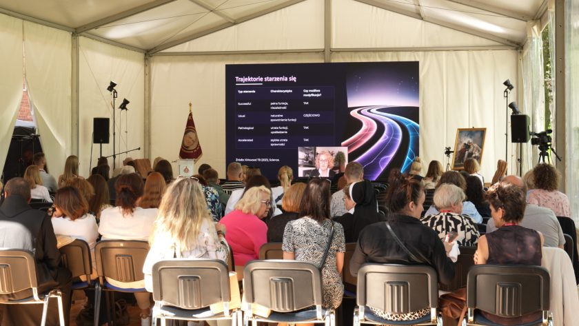 Uczestnicy siedzący w namiocie konferencyjnym, oglądający prezentację na dużym ekranie dotyczącą trajektorii starzenia się.