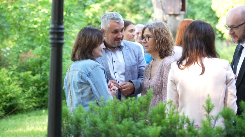 Grupa osób rozmawia na tle zieleni w ogrodzie. Na pierwszym planie trzyosobowa grupa: kobieta w dżinsowej kurtce, mężczyzna w jasnej marynarce i kobieta w okularach w różowej sukni. W tle inni uczestnicy spotkania i naturalne otoczenie.