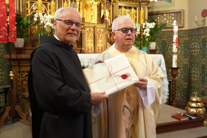 Dwaj duchowni, ubrani w uroczyste szaty liturgiczne, stoją przed ołtarzem trzymając zapakowany dokument przewiązany czerwonym sznurkiem z lakową pieczęcią. Tło stanowi bogato zdobiony ołtarz z kwiatami i świecami.