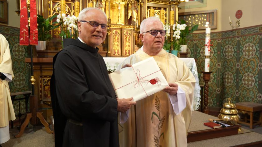 Dwaj duchowni, ubrani w uroczyste szaty liturgiczne, stoją przed ołtarzem trzymając zapakowany dokument przewiązany czerwonym sznurkiem z lakową pieczęcią. Tło stanowi bogato zdobiony ołtarz z kwiatami i świecami.