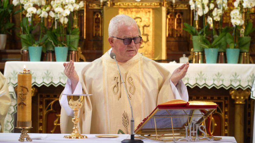 Starszy duchowny w okularach, ubrany w jasny ornat, celebruje mszę świętą. Jego dłonie są uniesione w geście modlitwy, przed nim kielich, mszał i mikrofon, a za nim kwiaty oraz ozdobny ołtarz z symbolami chrześcijańskimi.