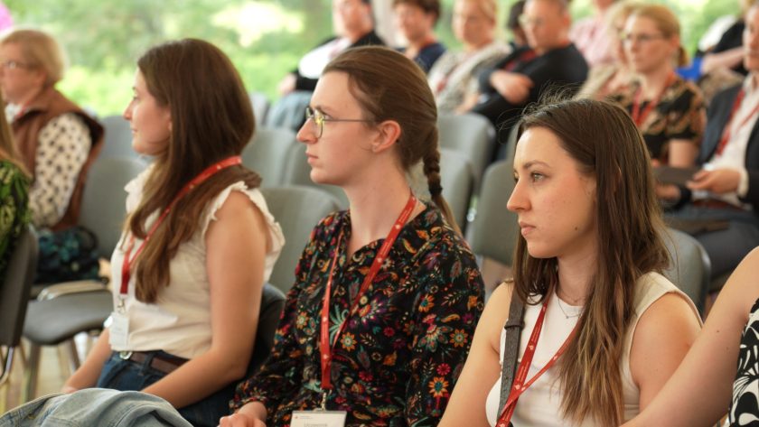 Trzy młode kobiety w pierwszym rzędzie sali konferencyjnej, skupione na prelekcji.