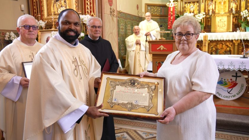 Uśmiechnięty duchowny i kobieta w białej sukience trzymają wspólnie dyplom oprawiony w ozdobną ramę. W tle duchowni i prezbiterium kościoła, w tym złocony ołtarz, świece i ornamenty sakralne.