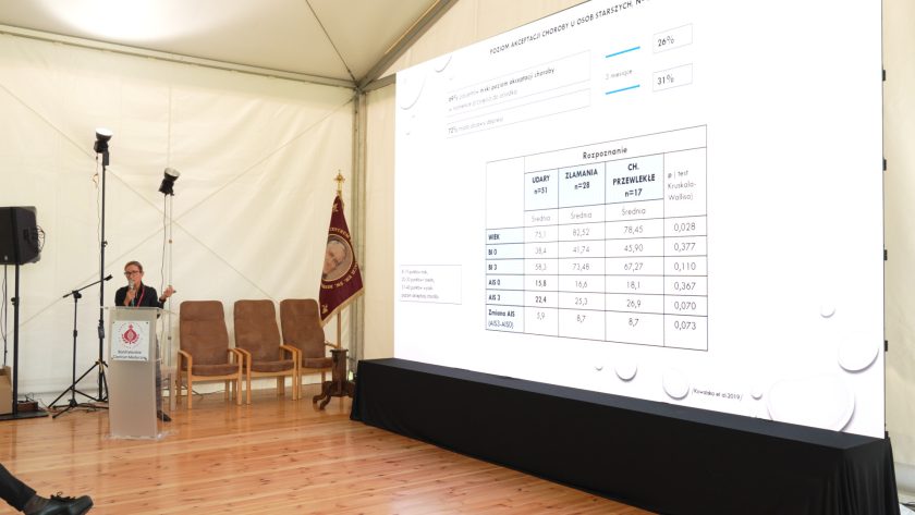 Widok ogólny sali konferencyjnej, prelegentka omawia dane z wyświetlonej prezentacji dotyczącej akceptacji choroby u osób starszych.