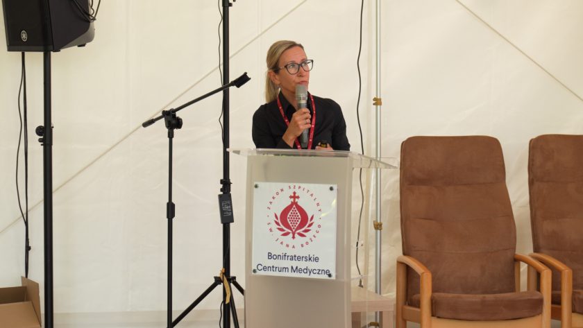Zbliżenie na prelegentkę przemawiającą zza podium z mikrofonem, w tle widoczne są krzesła dla panelistów.