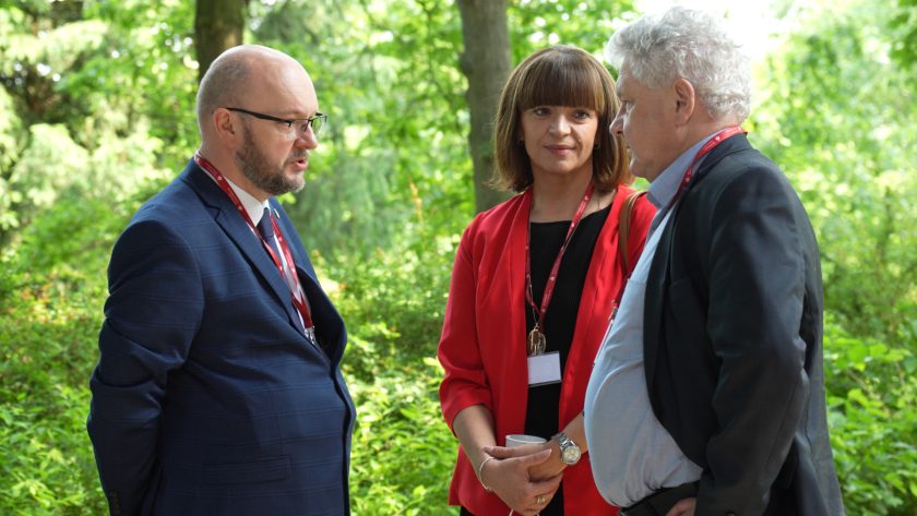 Trójka uczestników wydarzenia, w tym mężczyzna w garniturze i kobieta w czerwonym żakiecie, rozmawiających w zielonym otoczeniu plenerowym.