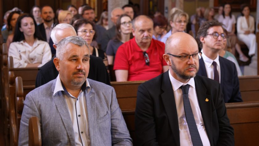 Mężczyźni w jasnej marynarce i garniturze siedzą w pierwszej ławce w świątyni, wsłuchani w treść uroczystości. W tle widać licznych uczestników nabożeństwa – kobiety i mężczyzn w skupieniu obserwujących przebieg wydarzenia.