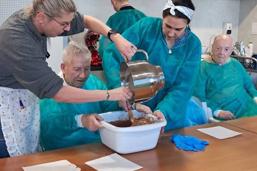 Zajęcia kulinarne w pracowni terapeutycznej. Starszy mężczyzna w fartuchu ochronnym siedzi przy stole i miesza w dużej misie brązową masę, podczas gdy dwie kobiety – opiekunka i uczestniczka zajęć – pomagają mu wlewając płyn z dużego metalowego dzbanka. W tle osoby w fartuchach ochronnych obserwują działania przy stole. Na pierwszym planie papierowe serwetki i niebieskie rękawiczki