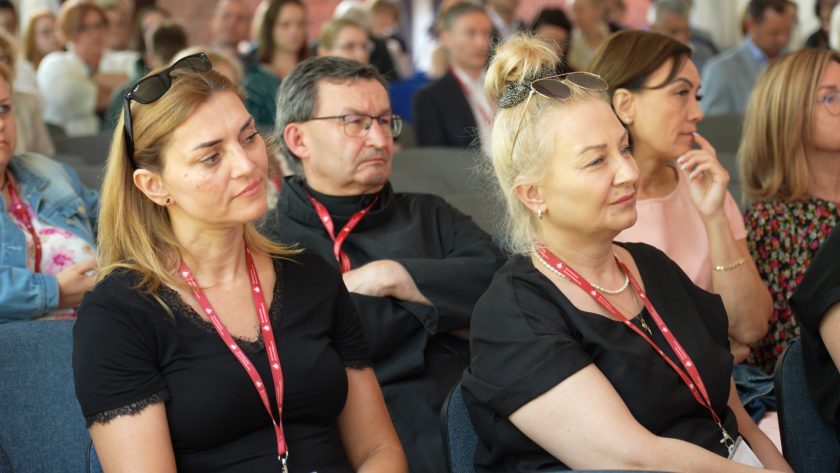 Kobiety ubrane w czarne bluzki oraz zakonnik w czarnym habicie wsłuchują się w wystąpienie, wśród skupionych uczestników spotkania.
