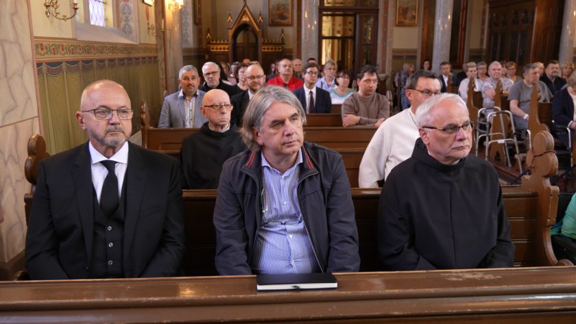 Trzech mężczyzn w garniturze i habitach siedzi w pierwszym rzędzie na tle pełnego kościoła.