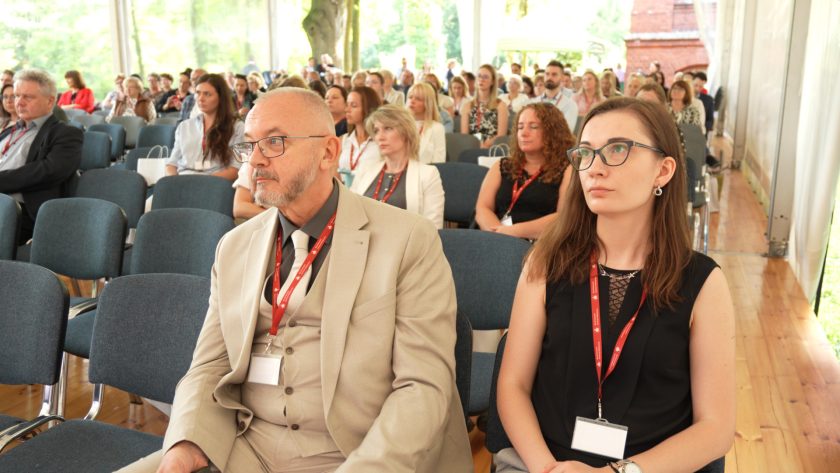 Dwoje uczestników w pierwszym rzędzie, starszy mężczyzna w beżowym garniturze i kobieta w czarnej sukience z czerwonym identyfikatorem, słuchają z uwagą prezentacji.