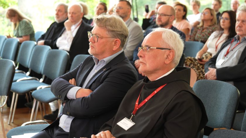 Mężczyzna w garniturze i brat zakonny w habicie siedzą obok siebie wśród licznych uczestników konferencji.
