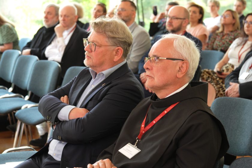 Mężczyzna w garniturze i brat zakonny w habicie siedzą obok siebie wśród licznych uczestników konferencji.