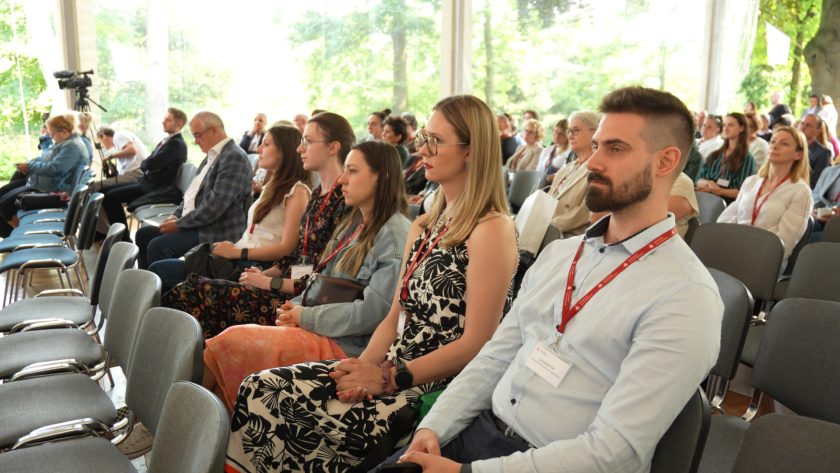 Szeroki kadr ukazujący rząd słuchaczy siedzących w pierwszym rzędzie. W tle widoczna licznie zgromadzona publiczność i duże przeszklenie namiotu konferencyjnego.