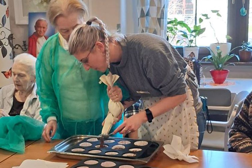 Dwie kobiety – pracownica i seniorka – wspólnie napełniają foremki na muffinki przygotowanym ciastem. Jedna z nich trzyma rękaw cukierniczy, druga pomaga przytrzymać formę. W tle widoczne są inne seniorki, dekoracje ścienne i parapet z kwiatami doniczkowymi, przez okno wpada dzienne światło. Atmosfera skupienia i zaangażowania