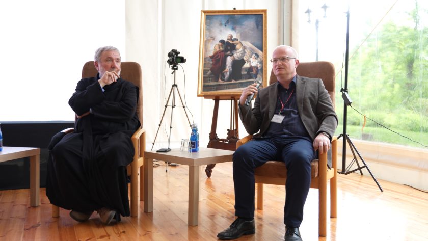 Dwóch mężczyzn prowadzi rozmowę na scenie, za ich plecami obraz z wizerunkiem świętego – moment refleksji i wymiany myśli.