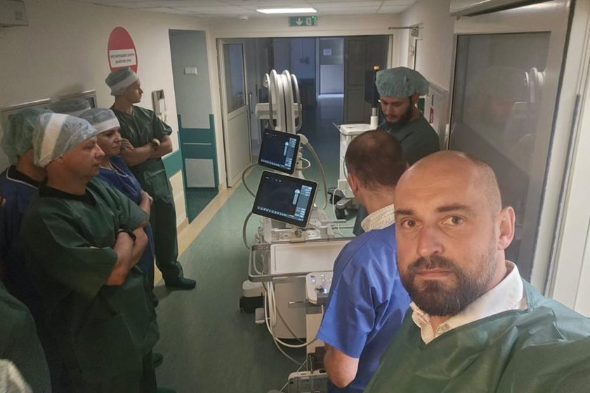 Zespół lekarzy i techników medycznych w strojach ochronnych podczas prezentacji aparatury do diagnostyki obrazowej na szpitalnym korytarzu.