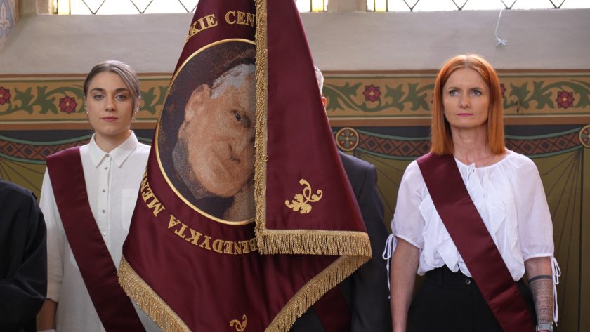 Dwie kobiety w białych koszulach i bordowych szarfach stojące obok sztandaru z wizerunkiem św. Jana Bożego. Znajdują się w kościele, na tle dekoracyjnych ścian i witraży. Sztandar zakrywa część postaci stojącej pomiędzy nimi.