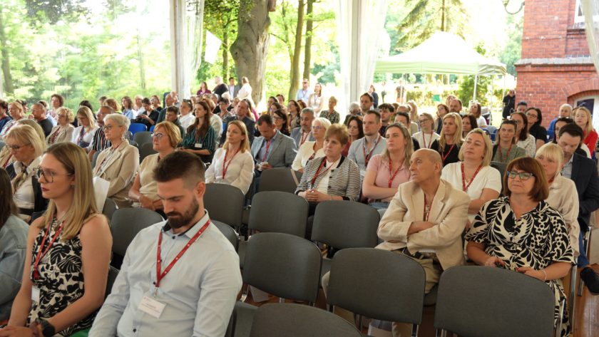 Szerokie ujęcie uczestników siedzących w rzędach podczas konferencji w namiocie. W tle drzewa i ceglany budynek, a uczestnicy skupieni na prezentacji.