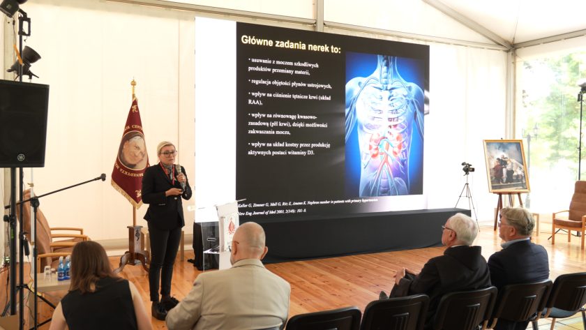 Prelegentka z mikrofonem prezentuje slajd o funkcjach nerek. Na ekranie widoczna grafika anatomiczna oraz lista zadań nerek.
