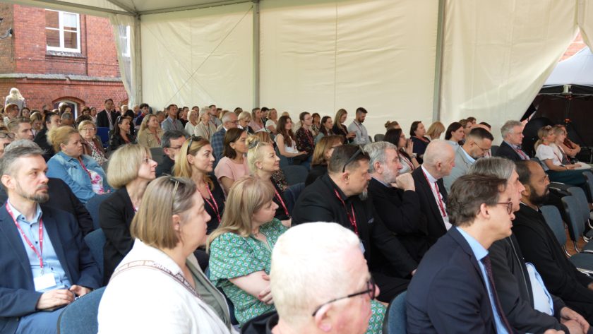 Ujęcie sali konferencyjnej z profilu. Liczni uczestnicy siedzą w rzędach i słuchają prezentacji, widoczna duża frekwencja.
