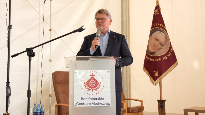 Mężczyzna w garniturze przemawia z mównicy z logo Bonifraterskiego Centrum Medycznego. Obok znajduje się sztandar zakonu.